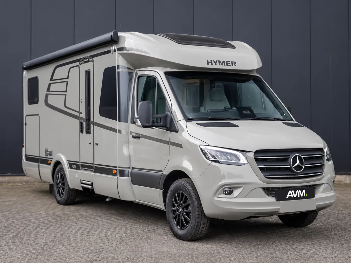 Beige Hymer-camper op Mercedes-Benz Sprinter-basis met AVM-kentekenplaat voor een donkere gevel